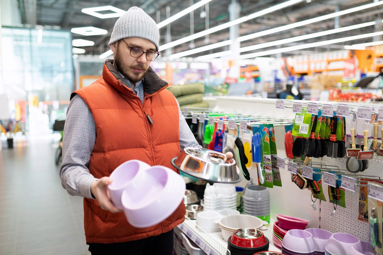 A man in an orange vest compares a lilac plastic dog bowl with a steel bowl in a pet store aisle