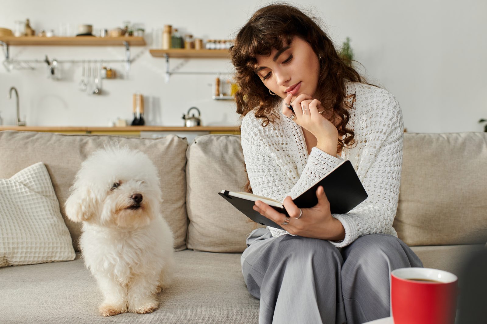A woman on a beige sofa holds an open notebook and rests her chin on her hand in thought while a white bichon sits beside her.