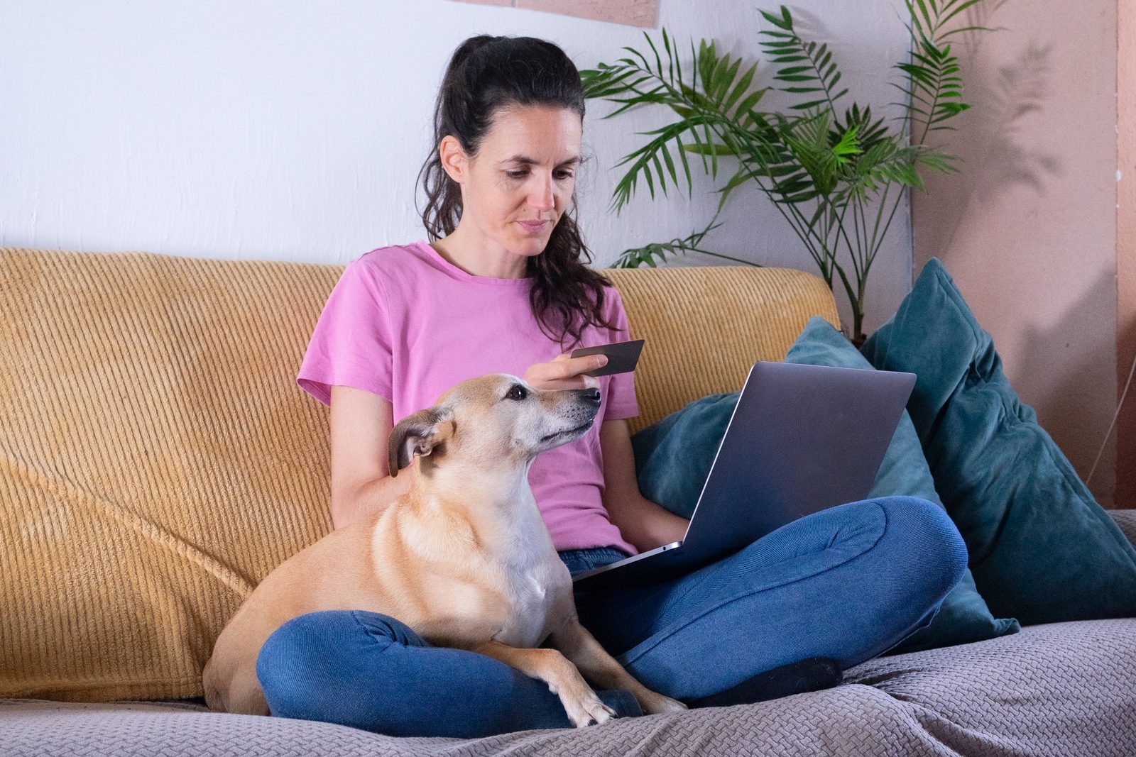 Woman in a pink T-shirt on a sofa holding a payment card and a laptop while a tan short-haired dog rests across her lap