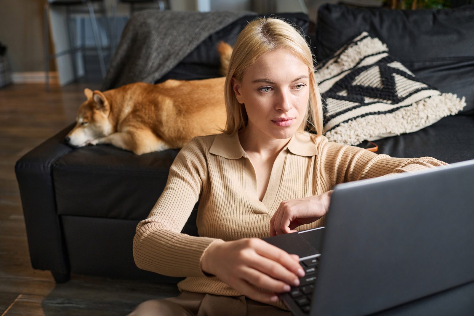 Woman seated on a living room floor typing on a laptop while a shiba inu naps on the black leather sofa behind her