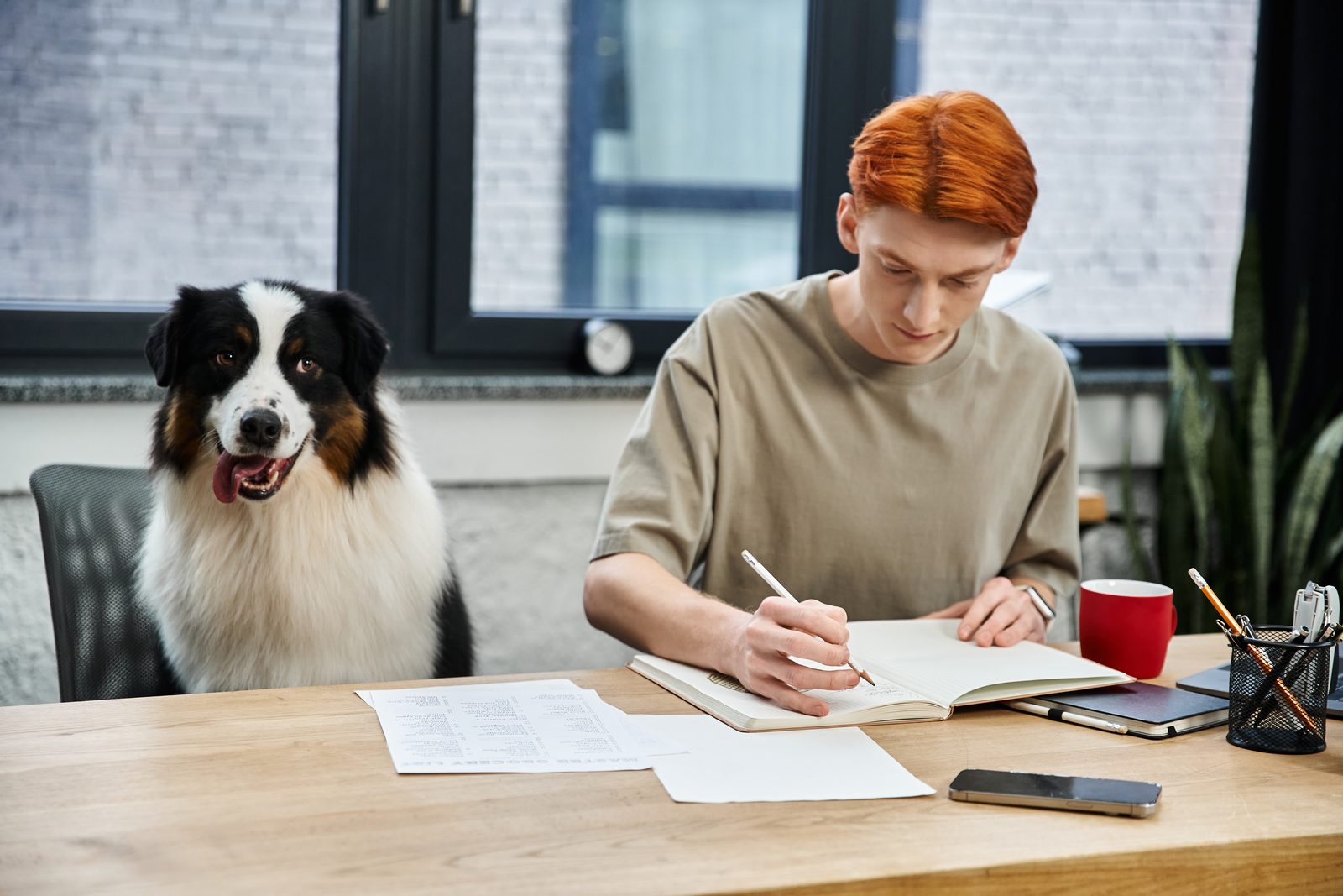 Man with red hair writing in a notebook at a wooden desk covered in papers while an Australian shepherd sits in the next chair