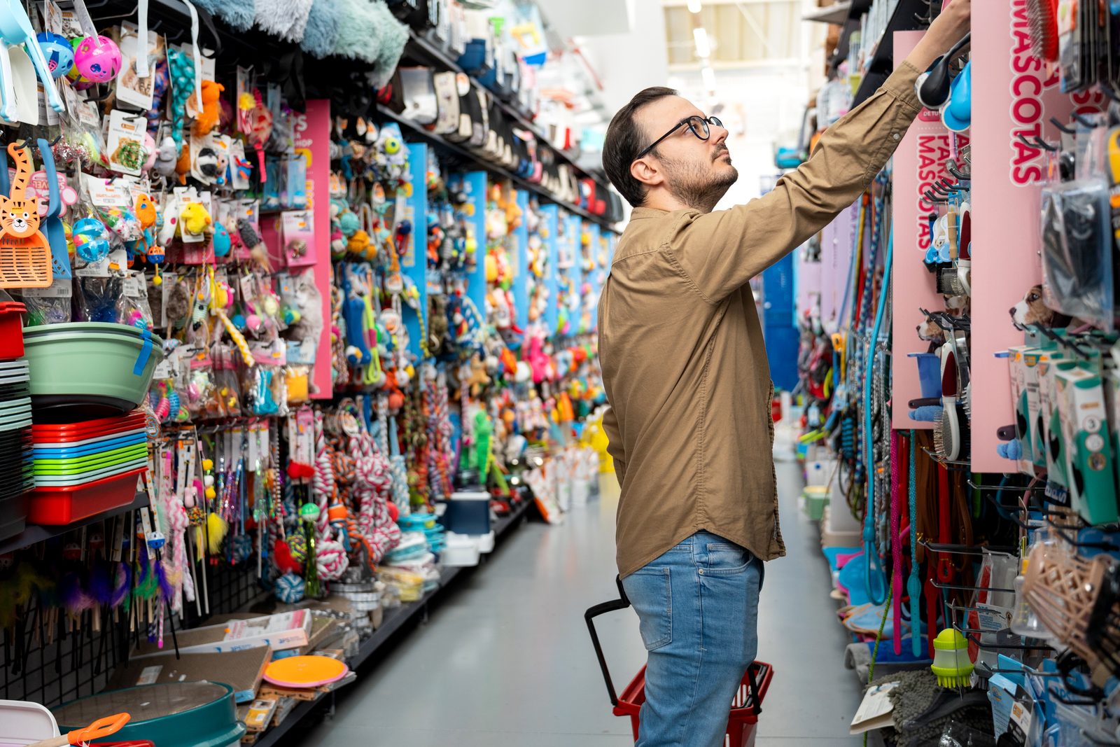 Man in glasses reaches for a product on a crowded pet store shelf lined with leashes, collars and toys, holding a red basket.