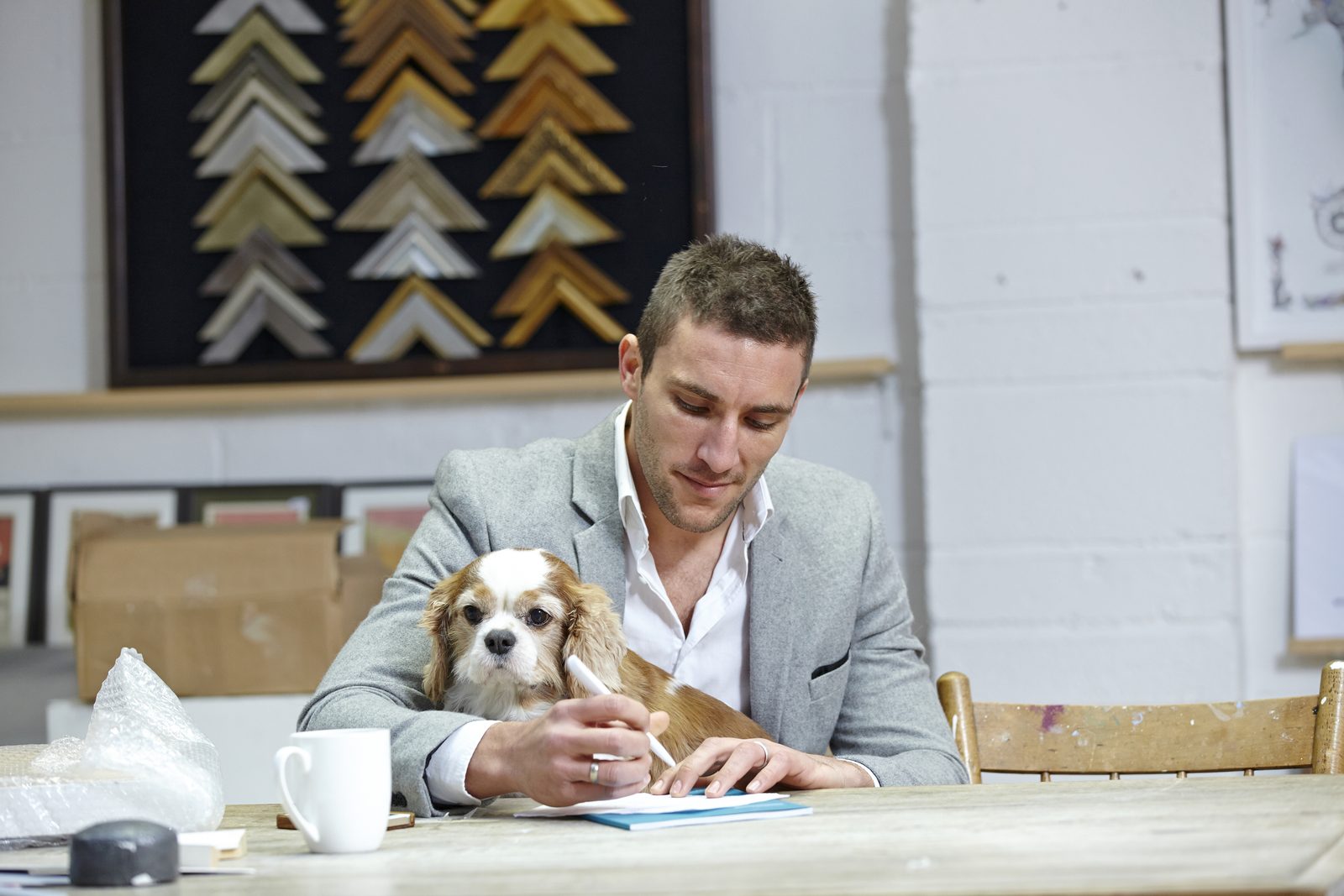 A man in a gray blazer writes on a sheet of paper at a wooden table while a Cavalier King Charles spaniel sits on his lap.